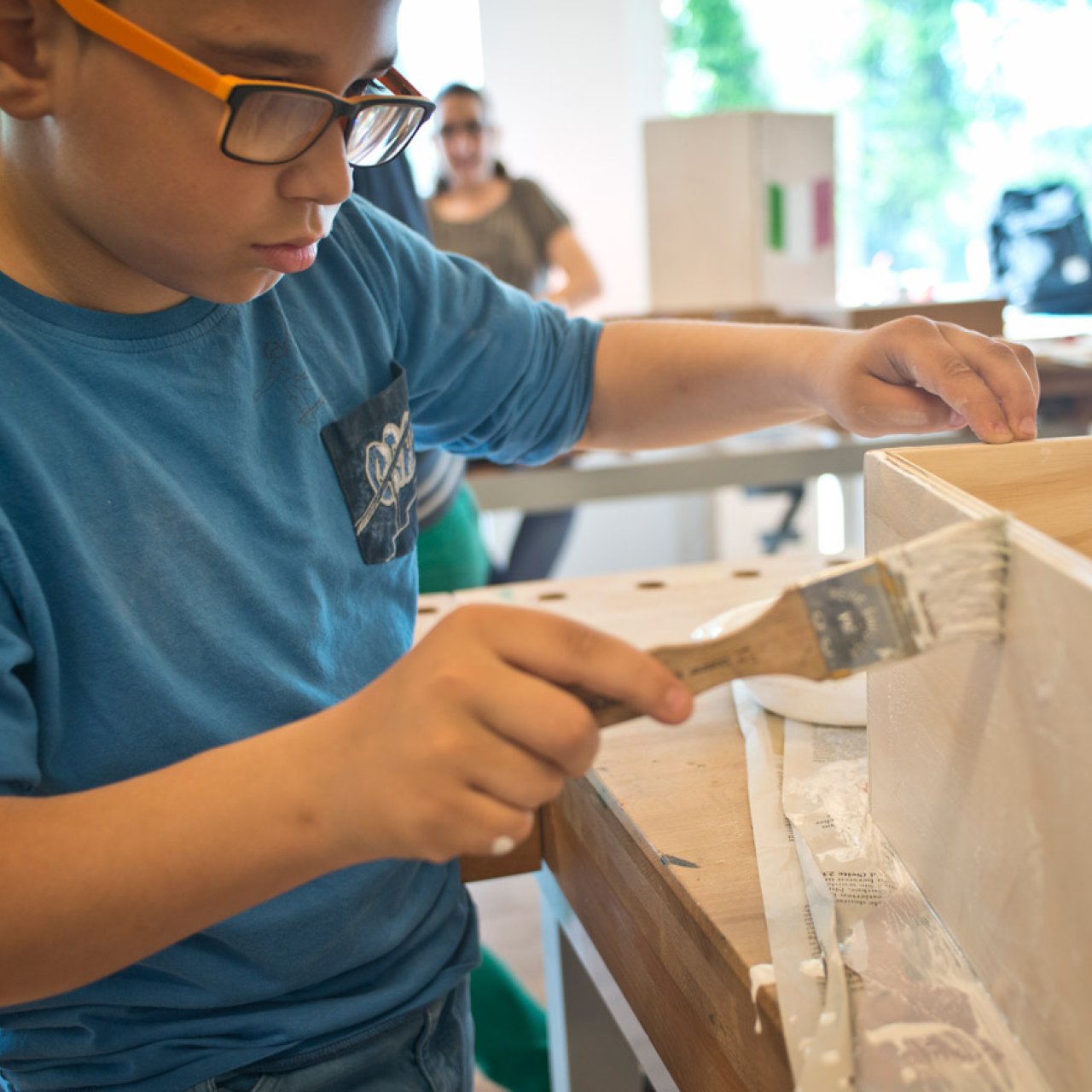 Aussenwohngruppe & Tagessonderschulen Bild Kind mit schwarz-oranger Brille malt eine Holzkiste weiss an